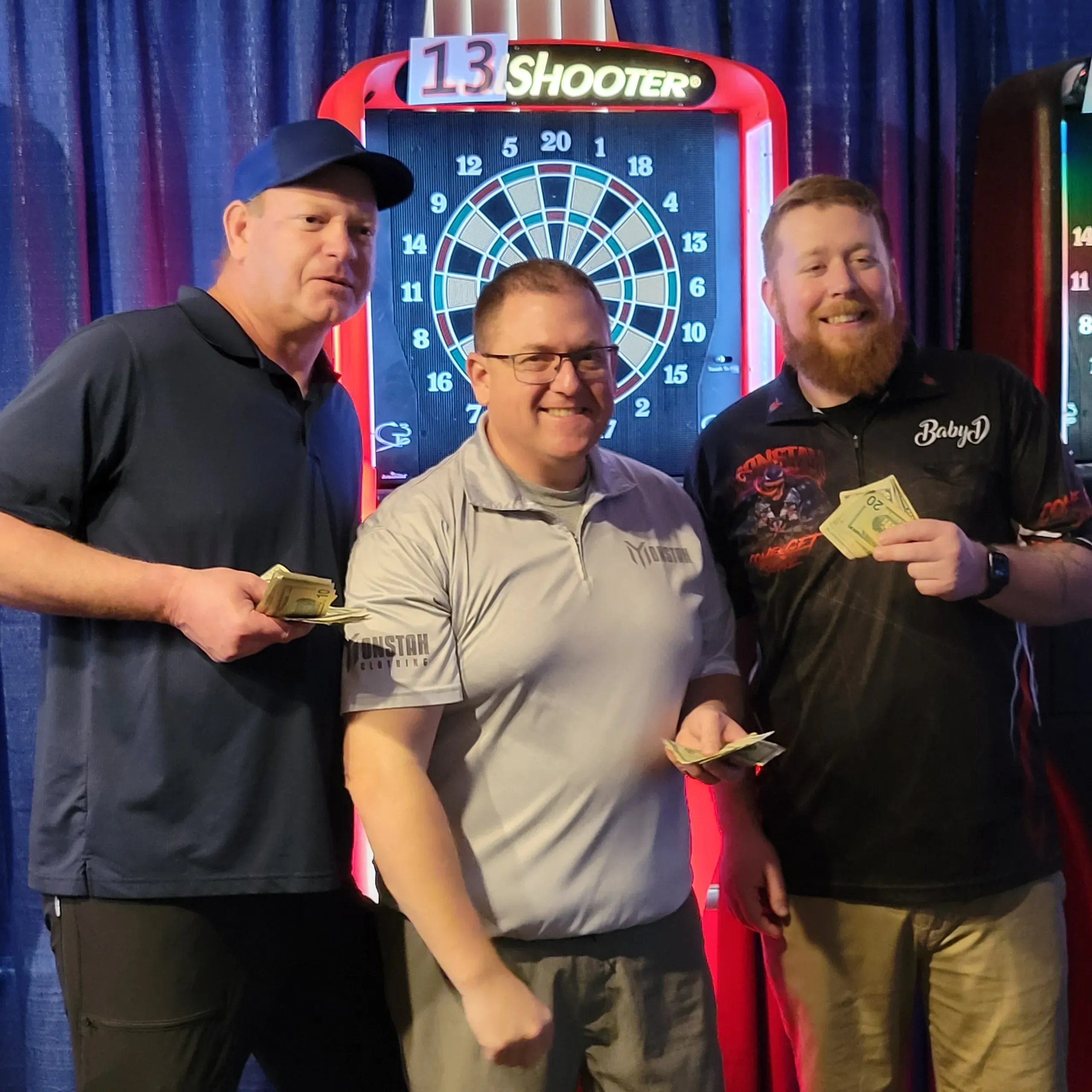 Three men holding money in front of dartboard.